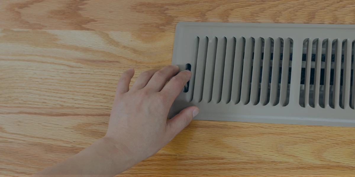 Close-up of a human hand (light skin) pressing a round, silver button on a rectangular metal floor vent. The vent is on a wooden floor.