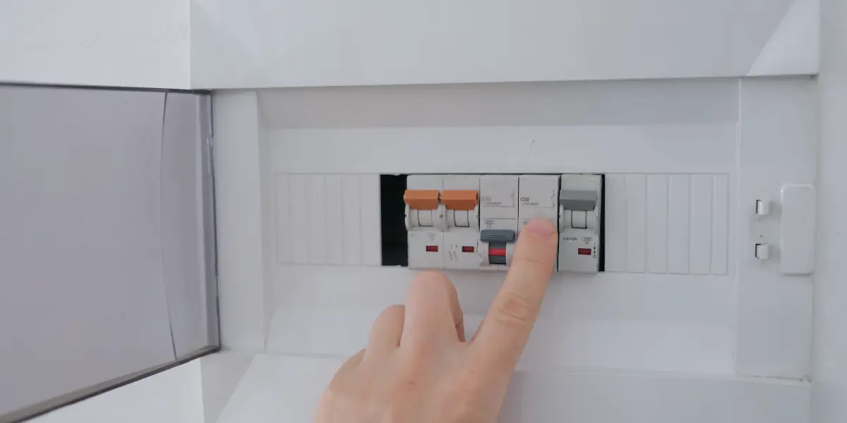 A close-up of a person's hand holding a flathead screwdriver, inserting it into the slot of a fuse box. The fuse box is made of white plastic and has several slots for fuses. There are warning labels on the fuse box.